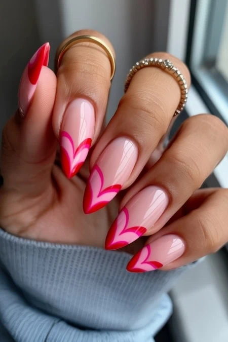 Bright pink and red heart nail art on well-manicured natural nails, with gold and beaded rings on fingers, showcasing stylish and creative nail design for Valentine's day.