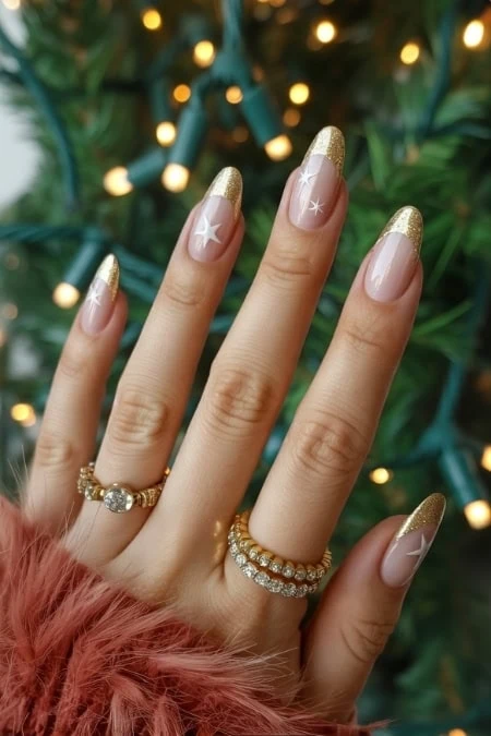 Elegant holiday-themed manicured nails with gold glitter tips and star designs, accessorized with rings, against a festive Christmas tree background.