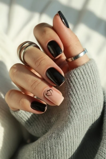 Black gel manicured nails with a heart tattoo on the ring finger, silver rings, and cozy gray sweater sleeve, showcasing elegant nail art and fashion accessories.