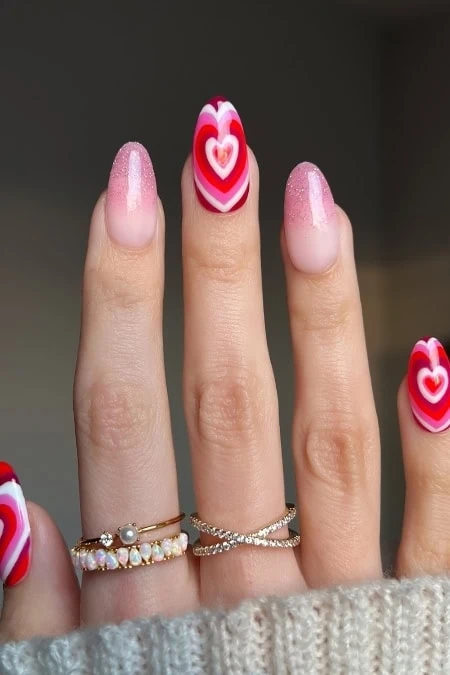 Vibrant Valentine's Day nail art with heart designs on ring finger, featuring pink and red shades, delicate rings, and a cozy sweater background. Perfect for romantic nail art inspiration.