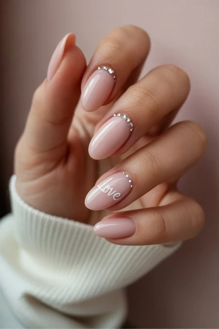 Elegant nude manicure with rhinestone accents and the word "Love" on the ring finger, perfect for beauty and nail art inspiration.