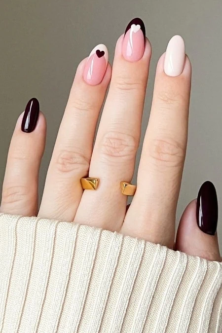 Elegant manicured nails with pink, white, and dark burgundy polish featuring heart accents, gold ring accessory, and cozy cream sweater sleeve, perfect for stylish everyday beauty.