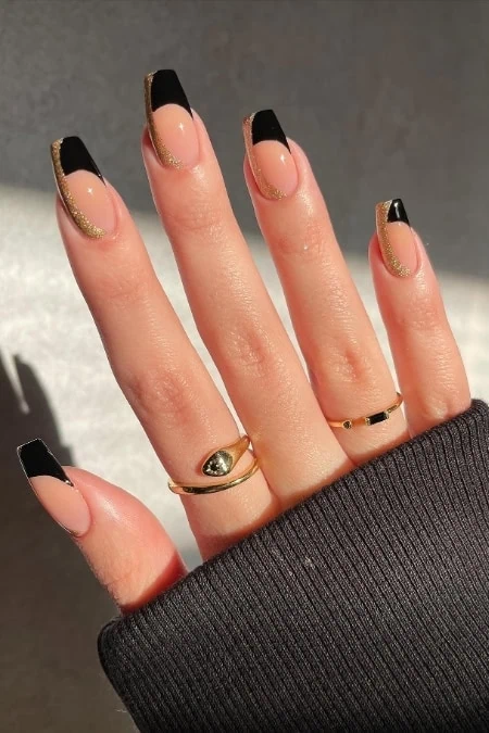 Glossy black and gold French tip manicure with glitter accents on natural nails, showcasing stylish nail art and elegant ring accessories for a trendy beauty look.