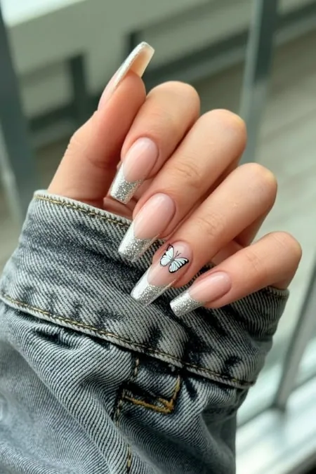 Elegant gel nail design with silver glitter French tips and a delicate butterfly accent on natural nail, perfect for modern nail art enthusiasts and beauty lovers.
