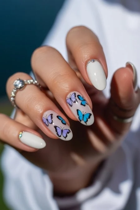 Elegant white Spanish nails with blue butterfly nail art and rhinestone accents, perfect for spring and summer fashion trends.