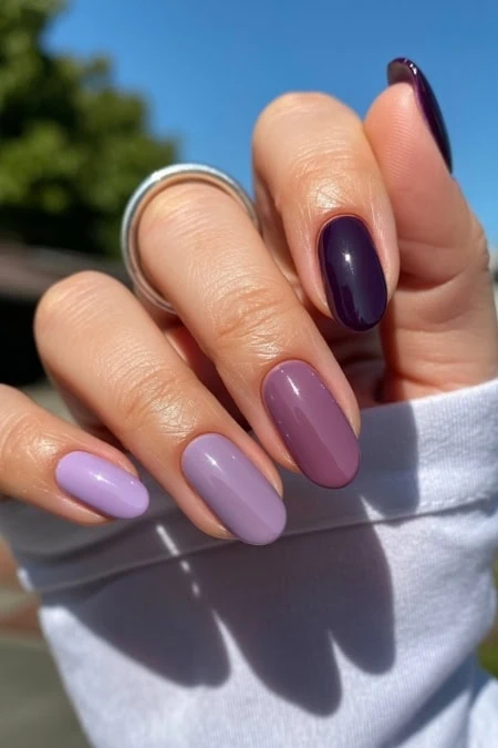 Close-up of a hand with beautifully manicured nails in purple and mauve shades, with a silver ring, outdoors on sunny day, emphasizing nail art and beauty.