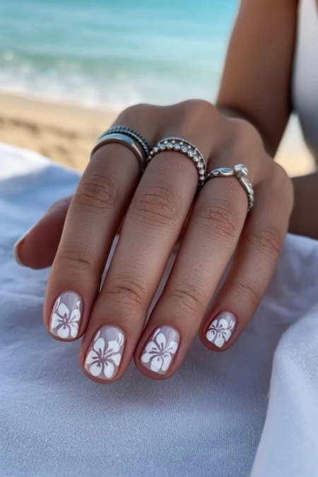 A woman’s hand displaying manicured nails with white Hawaiian-style hibiscus floral nail art, adorned with multiple silver rings, placed against a beach backdrop with sand and ocean waves.