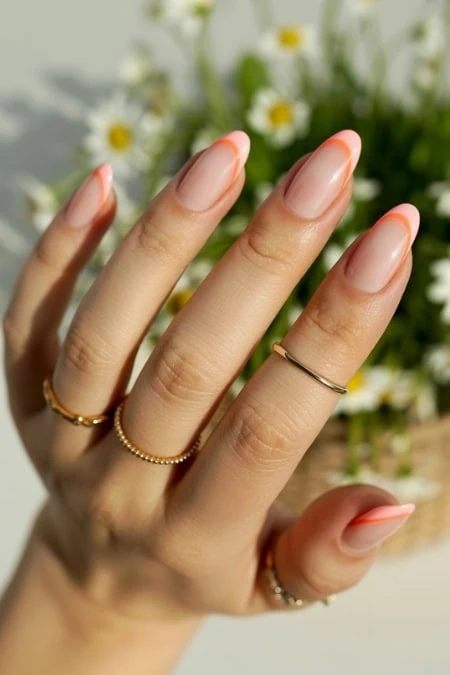 Beautiful manicured hand with nude and peach gradient gel nails, accessorized with gold rings, set against a background of white daisies, perfect for showcasing elegant nail art and self-care beauty routines.