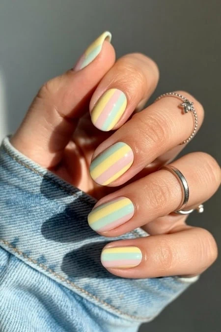 Pastel rainbow striped nail art with yellow, pink, blue, and mint green colors on short, well-manicured nails featuring rings for a stylish, feminine look.