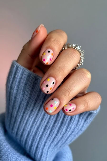 Colorful polka dot nail art with pink, yellow, black, blue, and orange dots on a nude base, accented by a silver ring, all on a person's hand with a cozy blue sweater. Perfect for showcasing creative nail design and trendy manicure ideas.