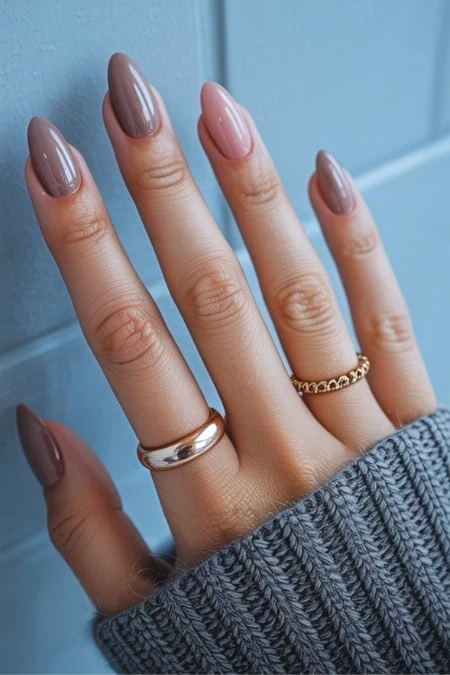 Well-manicured nails with nude and pink polish on a woman’s hand, wearing rings and a knitted sweater, against a blue background, showcasing professional nail art and beauty care.
