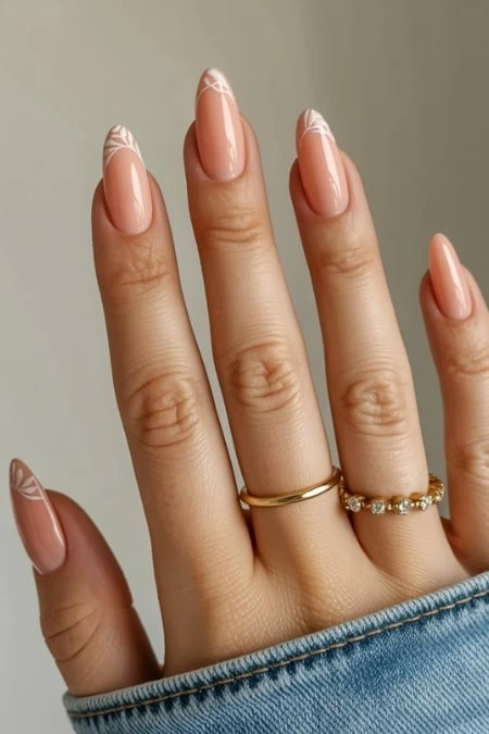 Soft pink almond-shaped manicured nails with delicate white nail art designs, accessorized with gold rings on a woman's hand, showcasing elegant nail art and jewelry.