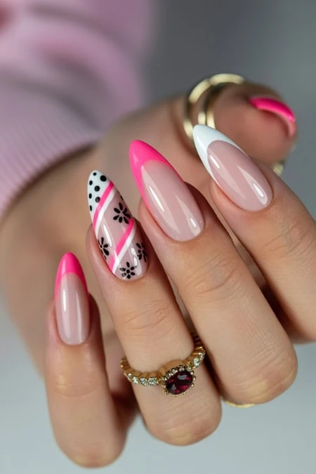 Vivid pink and white almond-shaped manicured nails with floral and polka dot designs, featuring a gold ring with a red gemstone, showcasing professional nail art and beauty care.