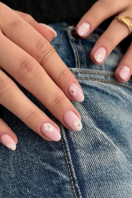 Soft pink nail polish with white floral designs on natural nails, paired with gold rings, resting on blue denim jeans, showcasing trendy nail art and fashion accessories for a stylish look.