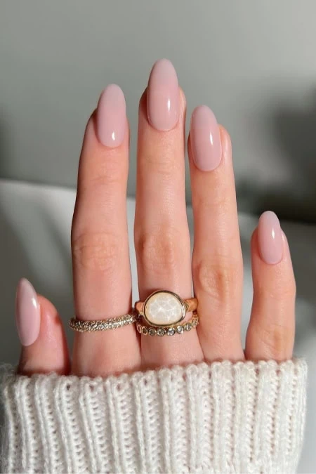 Soft pink, almond-shaped manicured nails with elegant gold rings featuring gemstones, held in front of a cozy knit sweater, showcasing stylish and sophisticated nail art perfect for beauty and fashion enthusiasts.