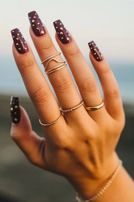 Vegan purple nail polish with gold stud accents on well-manicured nails, showcasing stylish jewelry including rings and bracelets for a trendy, feminine look.