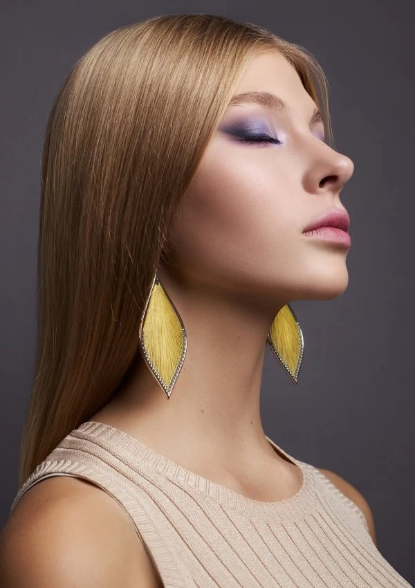 Blonde woman with purple eye makeup and dangling yellow earrings, close-up portrait, showcasing beauty and makeup skills for spring, gray background.