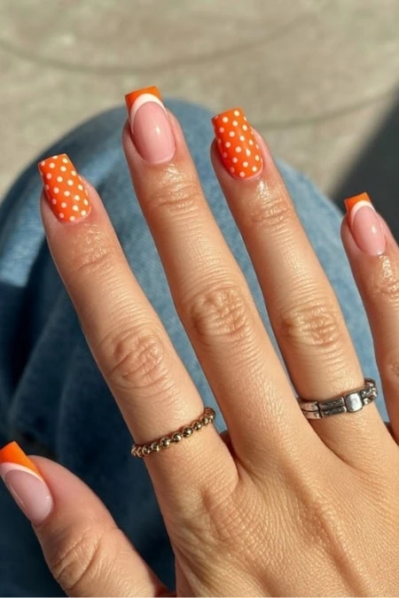 hand showcasing vibrant orange and white polka dot manicure with well-groomed square nails, featuring a silver ring and a delicate gold ring, against a blurred background.