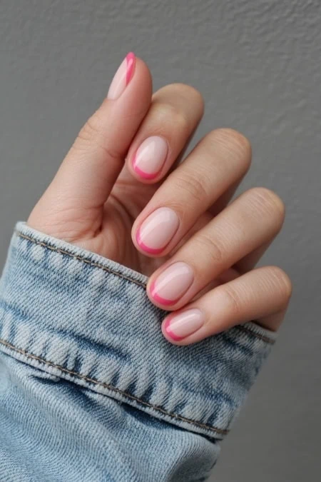 Woman's hand with a French manicure featuring soft pink tips and a natural nail base, wearing a light denim jacket sleeve against a neutral gray background.