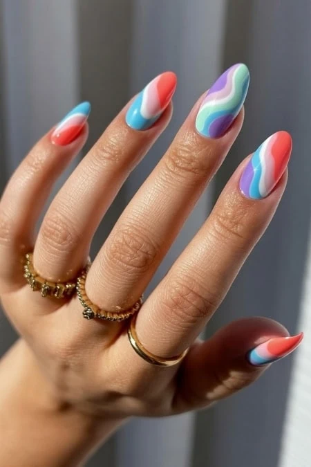 Vibrant, multicolored swirl nail design featuring shades of blue, purple, pink, and white on a well-manicured hand with gold rings, showcasing creative and trendy nail art for beauty and fashion enthu.