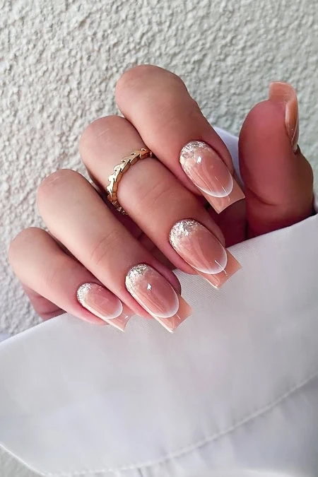 Elegant French manicure with glitter accents on the tips, featuring a gold ring on the finger, showcasing a sophisticated and stylish nail design perfect for special occasions or everyday elegance.