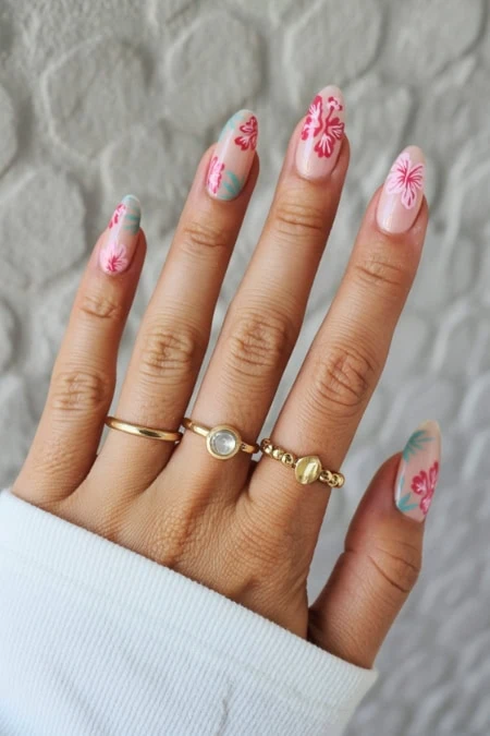 Beautiful hand with floral nail art featuring pink and green designs, accessorized with gold rings, against a textured white background.