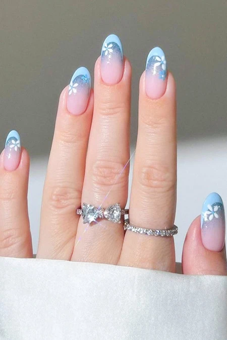 Blue and white floral nail art featuring a gradient design with daisy accents, complemented by silver rings on fingers, showcasing stylish and trendy manicure for beauty and fashion enthusiast.