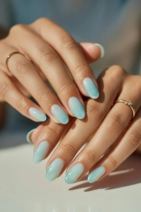 Close-up of a woman's hands showcasing a stylish soft aqua blue jelly manicure with well-groomed nails and elegant gold rings, emphasizing beauty and nail care.