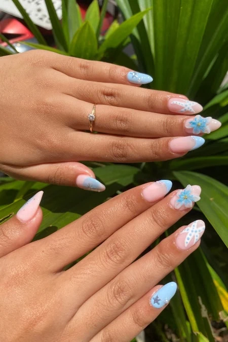 Beautiful hand with pastel blue and pink floral nail art, including 3D flowers and glitter accents, displayed against lush green foliage. The manicure highlights trendy, artistic nail designs suitable.