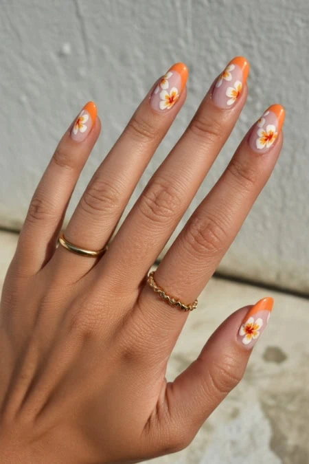 Elegant hand with floral nail art featuring orange tips and white flowers, showcasing professional manicure and jewelry, against a neutral background.