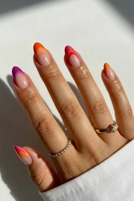 Brightly colored, stylish French manicure with vibrant orange, pink, and purple tips on well-groomed nails, adorned with elegant rings, against a neutral background.
