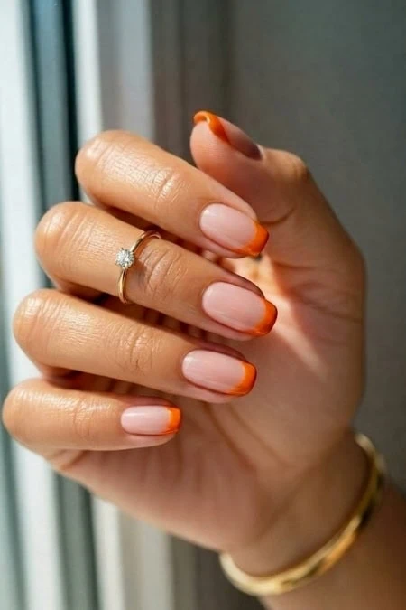 A woman’s hand with manicured nails featuring a natural pink base and orange tips, adorned with a diamond ring and gold bracelet, near a window.