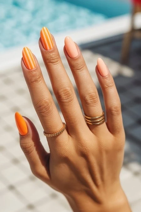 Elegant hand with manicured nails featuring orange and peach striped and solid nail polish, adorned with gold rings, against a blurred outdoor poolside background, showcasing beauty and self-care.