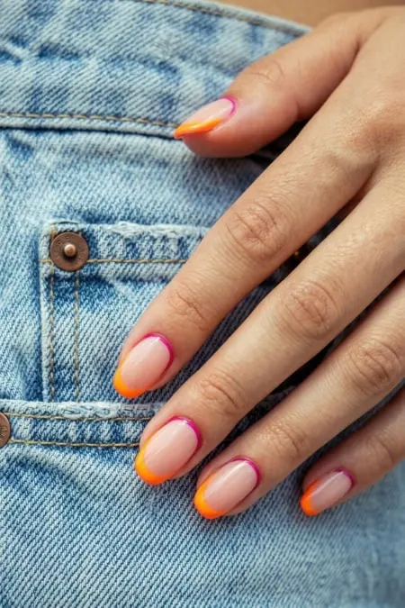 Brightly colored French manicure with orange tips on well-groomed nails resting on blue denim jeans, showcasing trendy nail art and casual fashion style.