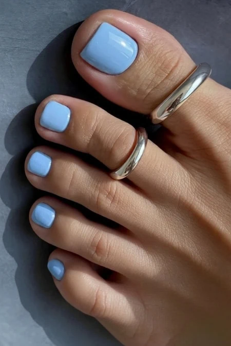 Elegant blue pedicure nails with a glossy finish, complemented by silver rings on fingers, showcasing a stylish and modern look for nail art and beauty enthusiasts.