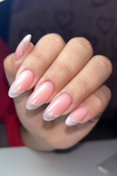 A detailed close-up of a woman's hand showcasing elegant, almond-shaped nails with a soft pink base and silver glitter tips, highlighting professional manicure and nail art.