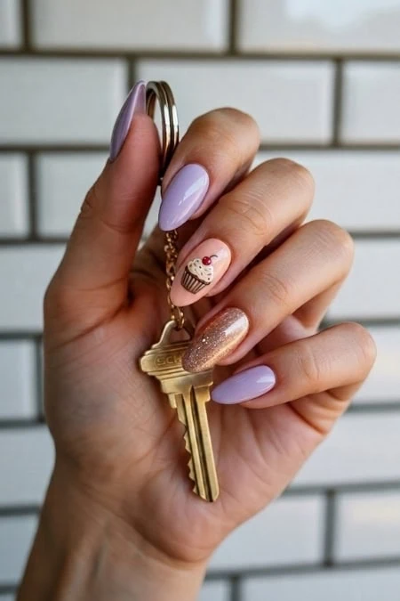 A key with a cupcake charm hanging from a hand with manicured nails, showcasing stylish nail art and jewelry, against a white brick wall background.