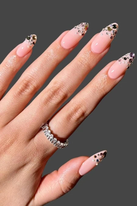 A detailed close-up of a woman's hand showcasing elegant, almond-shaped nails with a soft pink base and intricate rhinestone embellishments at the tips, featuring a sparkling ring on the ring finger.