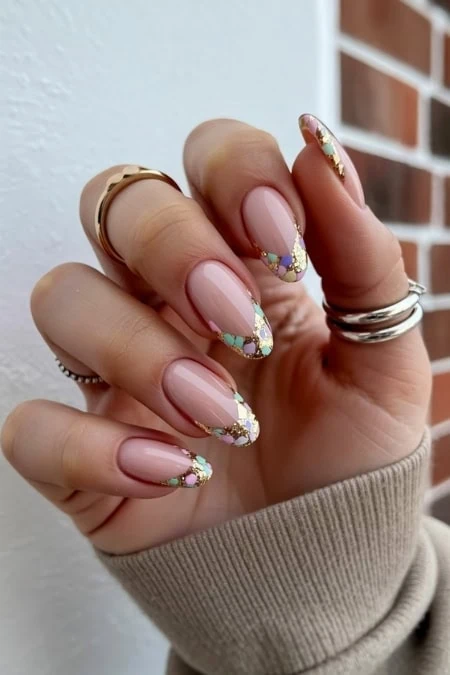 Elegant manicured nails with intricate multicolored gemstone nail art, featuring a soft pink base and decorative accents, held against a neutral background with a brick wall.