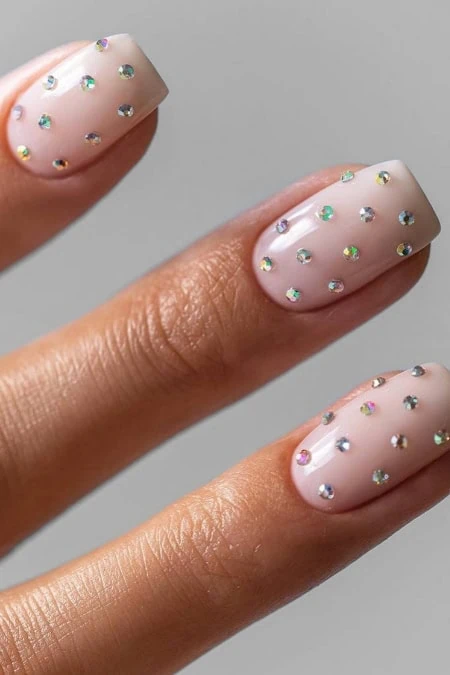 A detailed close-up of a woman's hand showcasing manicured nails with a soft pink base color and multicolored rhinestones, emphasizing nail art, beauty, and personal care.