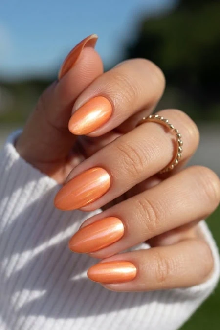 Elegant peach-colored almond-shaped manicured nails with a glossy finish, adorned with a delicate gold beaded ring, held against a blurred outdoor background.