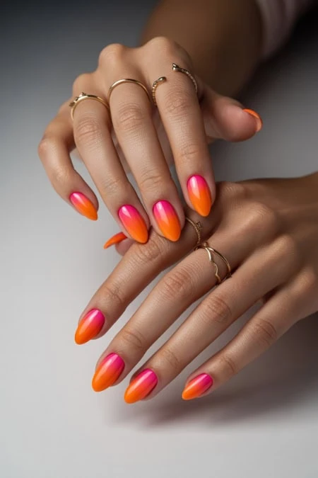 Brightly colored ombre nail art featuring pink and orange gradient on well-manicured hands with multiple gold rings, showcasing trendy and vibrant nail design.