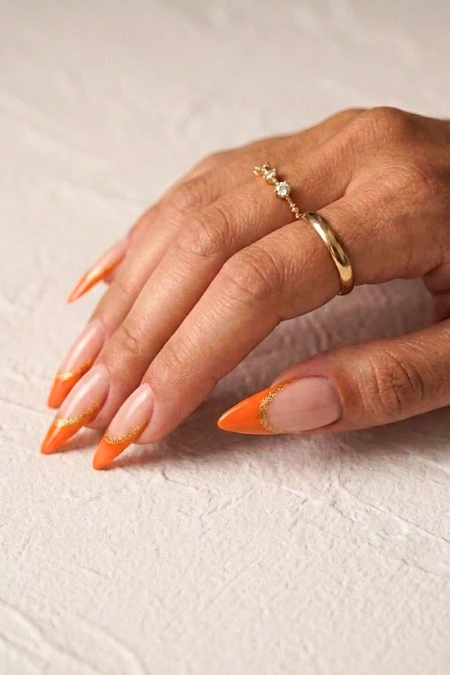 A woman’s hand with manicured nails featuring orange tips, adorned with a gold ring and a delicate diamond ring, resting on a textured light-colored surface.