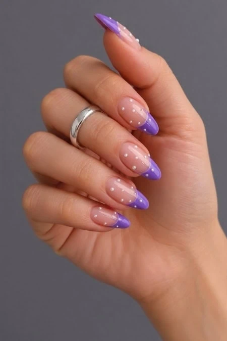 A detailed close-up of a woman's hand showcasing elegant, long almond-shaped nails with a purple French manicure and white polka dots, against a neutral gray background.