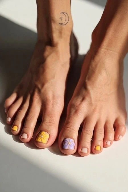 A close-up of well-groomed feet featuring a beach-inspired pedicure with yellow and purple nail polish, floral designs, and a small moon and star tattoo on the ankle, perfect for summer skincare and b.