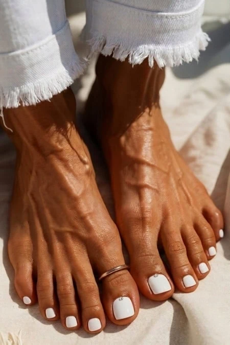 Feet with white pedicure on sandy beach, casual style, relaxed summer vibe, natural skin tone, and frayed denim jeans.