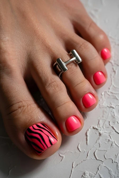 Pedicure with pink and zebra nail art, featuring a silver toe ring on the second toe, on textured white background.