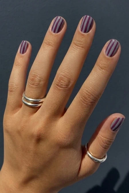 Close-up of a hand with striped purple and white nail polish, wearing silver rings on the index and middle fingers, against a dark background.