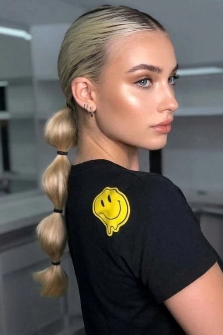 A woman with platinum blonde hair styled in a bubble braid, wearing a black t-shirt with a yellow smiley face graphic, in a modern indoor setting.