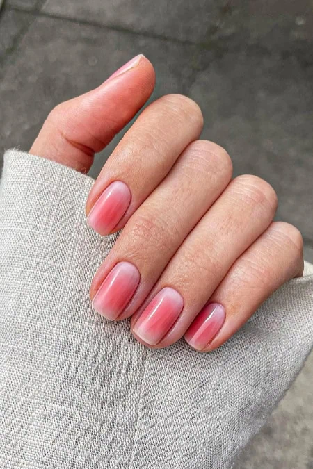 Close-up of a hand with well-manicured nails featuring a pink ombre nail design, held against a neutral background.